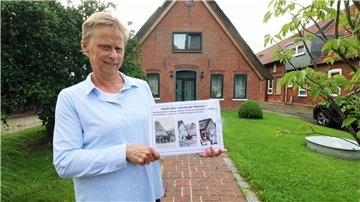 Karla Mombeck hält eine Infotafel vor einem Haus mit jüdischer Geschichte in Sandstedt