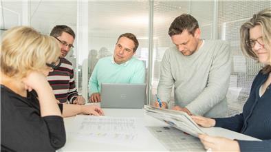 Männer und Frauen stehen an einem Tisch und konferieren.