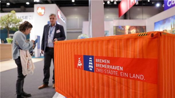Ein Mann und eine Frau stehen neben einem Mini-Container an einem Messestand.