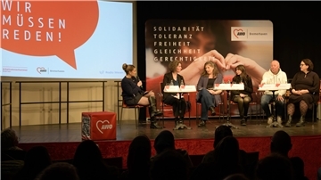 Podiumsdiskussion auf einer Bühne mit sechs Teilnehmenden und einer Moderatorin bei der Veranstaltung „Wir müssen reden!“ von AWO Bremerhaven und der Arbeitnehmerkammer. Im Hintergrund sind ein großes Veranstaltungsbanner und ein AWO-Würfel zu sehen.