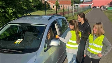 Zwei Schülerinnen in Warnwesten überreichen einer Autofahrerin eine Zitrone, begleitet von einer Polizistin, im Rahmen einer Verkehrssicherheitsaktion.