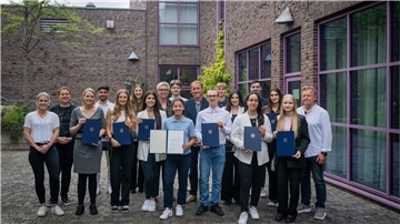 Gruppenfoto von vielen Menschen vor einem Gebäude. Einige halten Urkunden und blaue Mappen in den Händen. Stadtrat Hauke Hilz überreichte im Historischen Museum Bremerhaven 15 Lehramtsstudierenden feierlich die Stipendienurkunden.