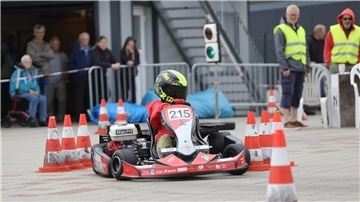 Ein Kartfahrer fährt auf einer Strecke zwischen Pylonen. Im Hintergrund sind Helfer in Warnweste und einige Zuschauer zu sehen.