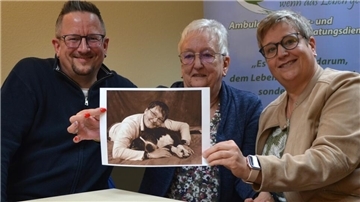 Ein Mann und zwei Frauen. Die Frauen halten ein Foto hoch.