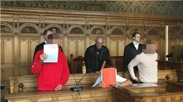 Drei Angeklagte auf der Bank, drei Anwälte stehen hinter ihnen.