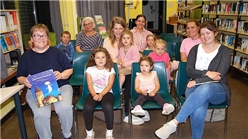 Mehrere Kinder und Erwachsene sitzen in einer gemütlichen Runde in einer Bücherei und blicken lächelnd in die Kamera. Links vorne hält eine Frau das Buch „Das kleine Gespenst“ von Otfried Preußler in der Hand. Im Hintergrund stehen Bücherregale mit zahlreichen Büchern.