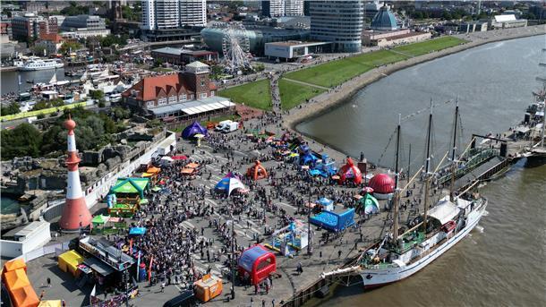 **ALT-Text:**
Luftaufnahme vom Willy-Brandt-Platz in Bremerhaven während eines Großevents mit bunten Pavillons, Aktionsflächen und Menschenmengen am Weserufer.