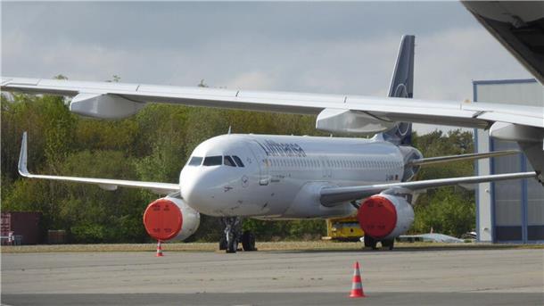 Ein geparktes Passagierflugzeug der Lufthansa, ein Airbus A320, steht auf einer Flughafenterrasse. Die Triebwerke sind mit roten Schutzabdeckungen versehen, und ein orange-weißer Verkehrskegel ist im Vordergrund sichtbar. Im Hintergrund sind Bäume und ein Hangar zu erkennen.