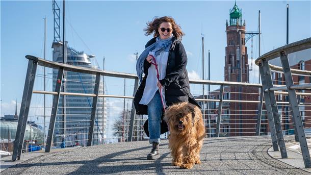 Eine Frau mit einem Briard-Hund lacht in die Kamera. Im Hintergrund sieht man das Atlantic Hotel Sail City in Bremerhaven.