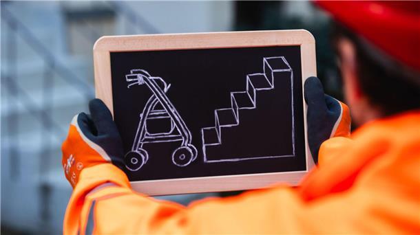 Ein Mann hält ein Schild mit einem Rollator vor einer Treppe.