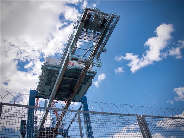 Wer nach links blickt, entdeckt hinter einem Zaun große Kräne, Container und riesige Autolandschaften.