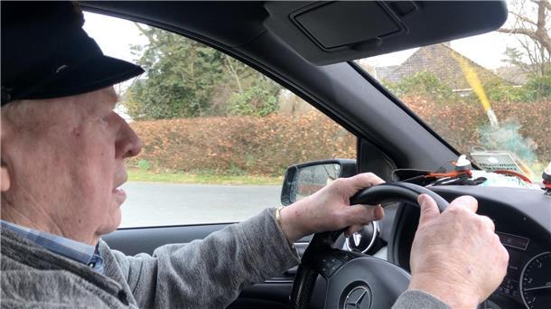 Älterer Mann mit schwarzer Mütze lehnt sich auf die Fahrertür seines schwarzen PKW.