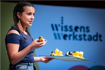 Eine junge Frau hält kleines Gummienten-Spielzeug in der Hand und präsentiert ein Tablett mit mehreren Gummienten-Spielzeugen vor einem Wissens Werkstadt-Schild."