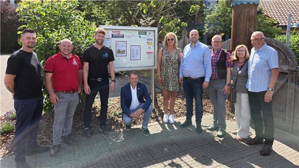 Hüpfburg und Schaukasten für Kita Nimmerland gespendet Eine Gruppe von Sponsoren und Organisatoren steht bei sonnigem Wetter vor dem neuen Informationsschaukasten des EWW Kindergartens Nimmerland.