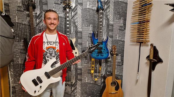 Ein junger Mann steht mit einer Gitarre in der Hand vor einer Wand, an der weitere Gitarren hängen, und lächelt in die Kamera.