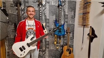 Ein junger Mann steht mit einer Gitarre in der Hand vor einer Wand, an der weitere Gitarren hängen, und lächelt in die Kamera.