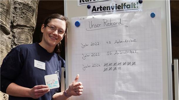 Eine Mitarbeiterin des Zoos am Meer steht lächelnd vor einer Tafel mit dem Titel „Unser Rekord“ und zeigt auf eine Liste gesammelter „Artendeckel“ der Jahre 2022 bis 2024.