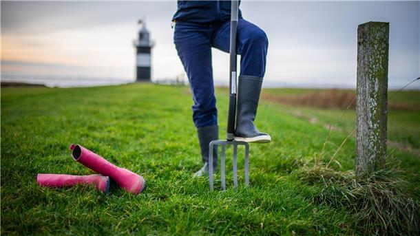 Eine Person trägt blaue Gummistiefel, daneben liegen pinke Stiefel im Gras. Im Hintergrund sieht man einen schwarz-weiß gestreiften Leuchtturm.