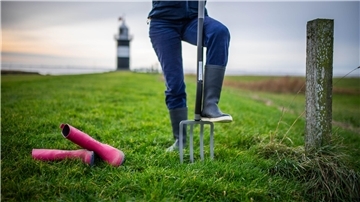 Eine Person trägt blaue Gummistiefel, daneben liegen pinke Stiefel im Gras. Im Hintergrund sieht man einen schwarz-weiß gestreiften Leuchtturm.