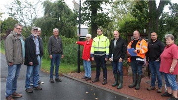 Eine Gruppe von zehn Personen steht bei bedecktem Wetter am Straßenrand in Rechtenfleth, teils auf Gehweg, teils auf der Fahrbahn. Einige tragen wetterfeste Kleidung und Gummistiefel. Im Hintergrund sind Bäume und ein Graben zu sehen.