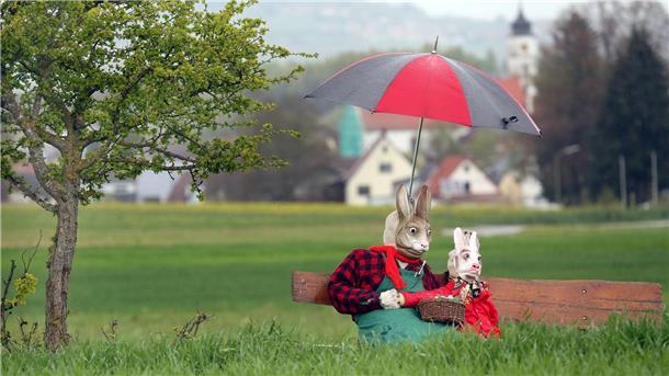 Zwei als Osterhasen dekorierte Strohpuppen sitzen mit einem großen Regenschirm an einem Feld.