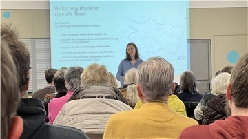 Im Hintergrund steht eine Frau mit langen dunkelblonden Haaren und im blauen Hemd. Sie hält einen Vortrag. Im Hintergrund ist eine Präsentation zu sehen. Im Vordergrund sitzen Menschen.