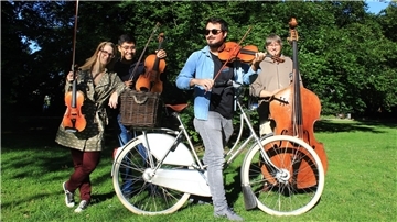 Vier Musiker stehen auf einer einer Wiese. Einer sitzt auf einem Fahrrad. Alle haben Instrumente dabei. 